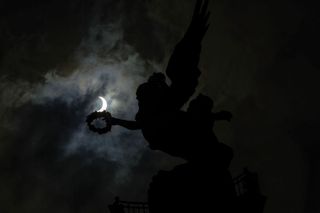 the sun looks like a crescent moon behind a silhouetted statue.