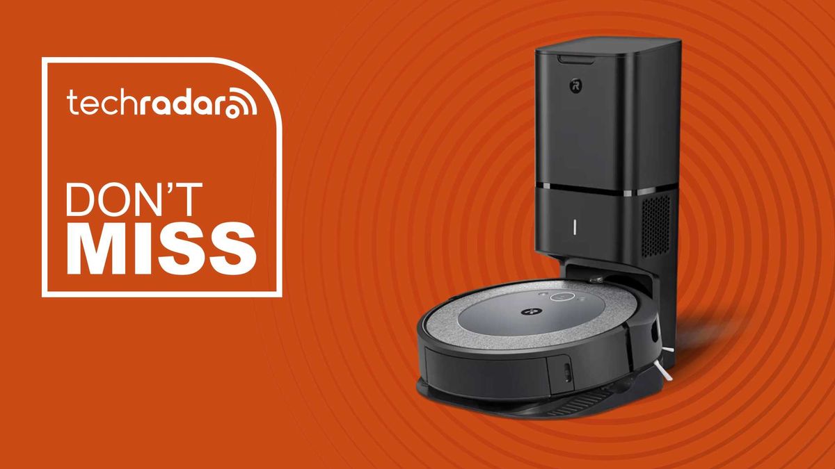 A Roomba robot vacuum sits on its charging station in front of an orange background. A sign reads &quot;Don&#039;t Miss.&quot;