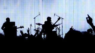 A band playing live against a bright bank of lights, with the crowd in front of them