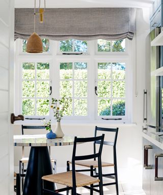 Large white framed windows with a grey roman blind hanging at the top in a white painted dining room. Black framed dining chairs around a round marble topped kitchen table.