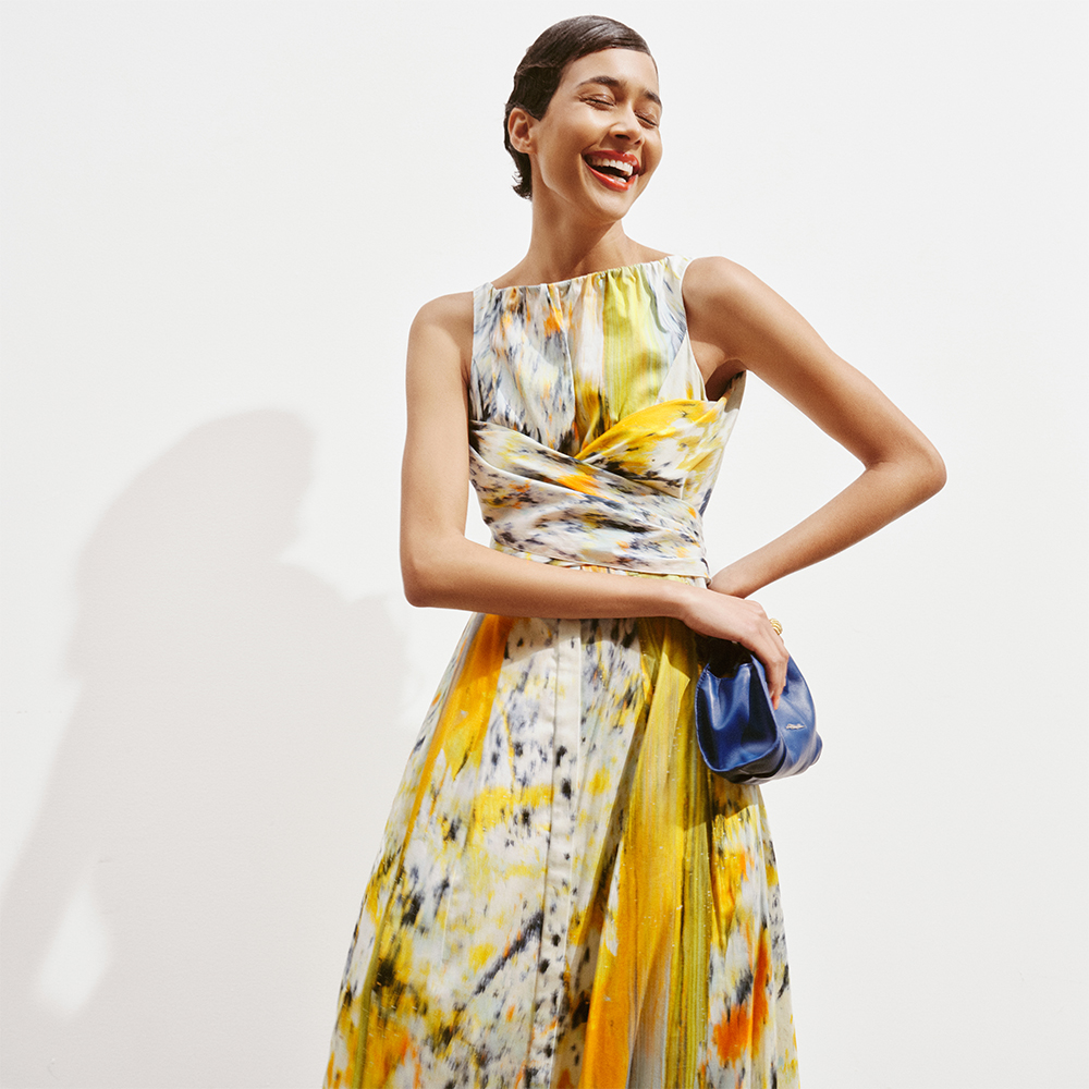Woman laughing wearing printed colourful dress from The Outnet and holding blue bag, with white background.
