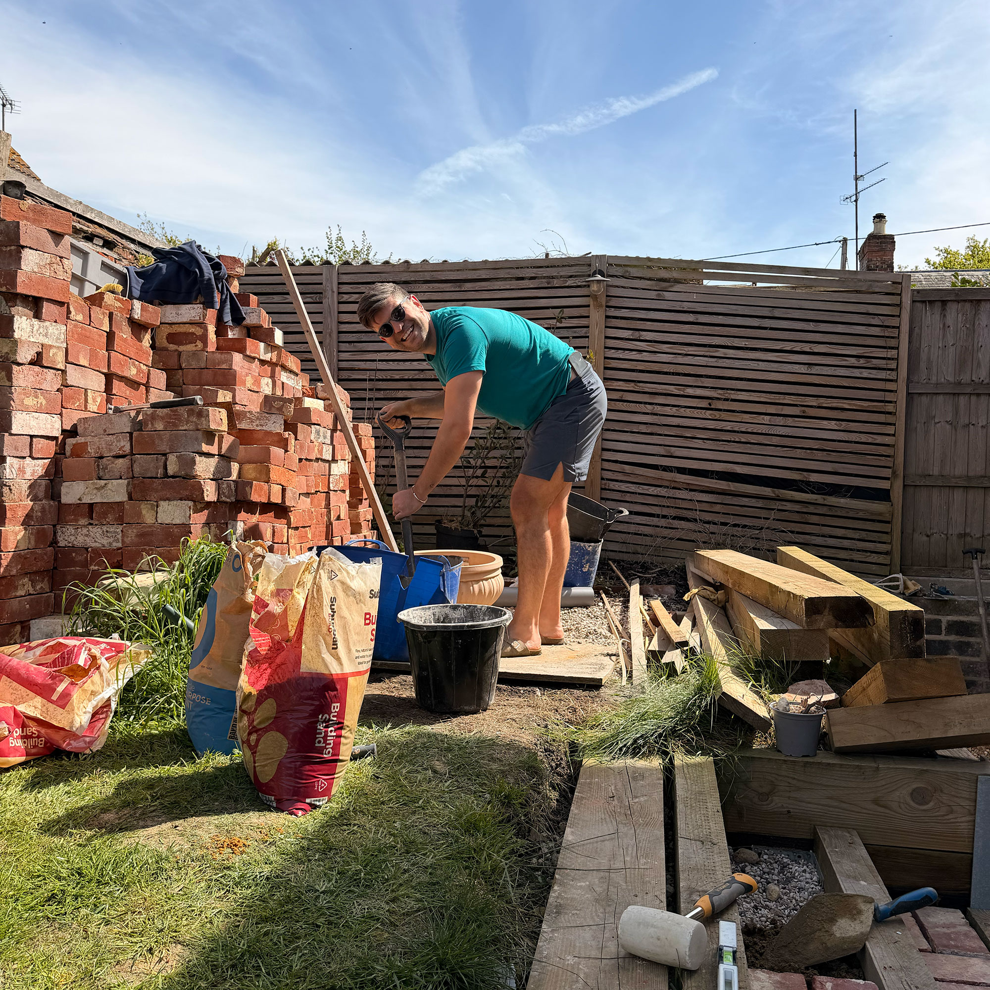 Man in garden beside pots, large piles of bricks, timber and bags of compost