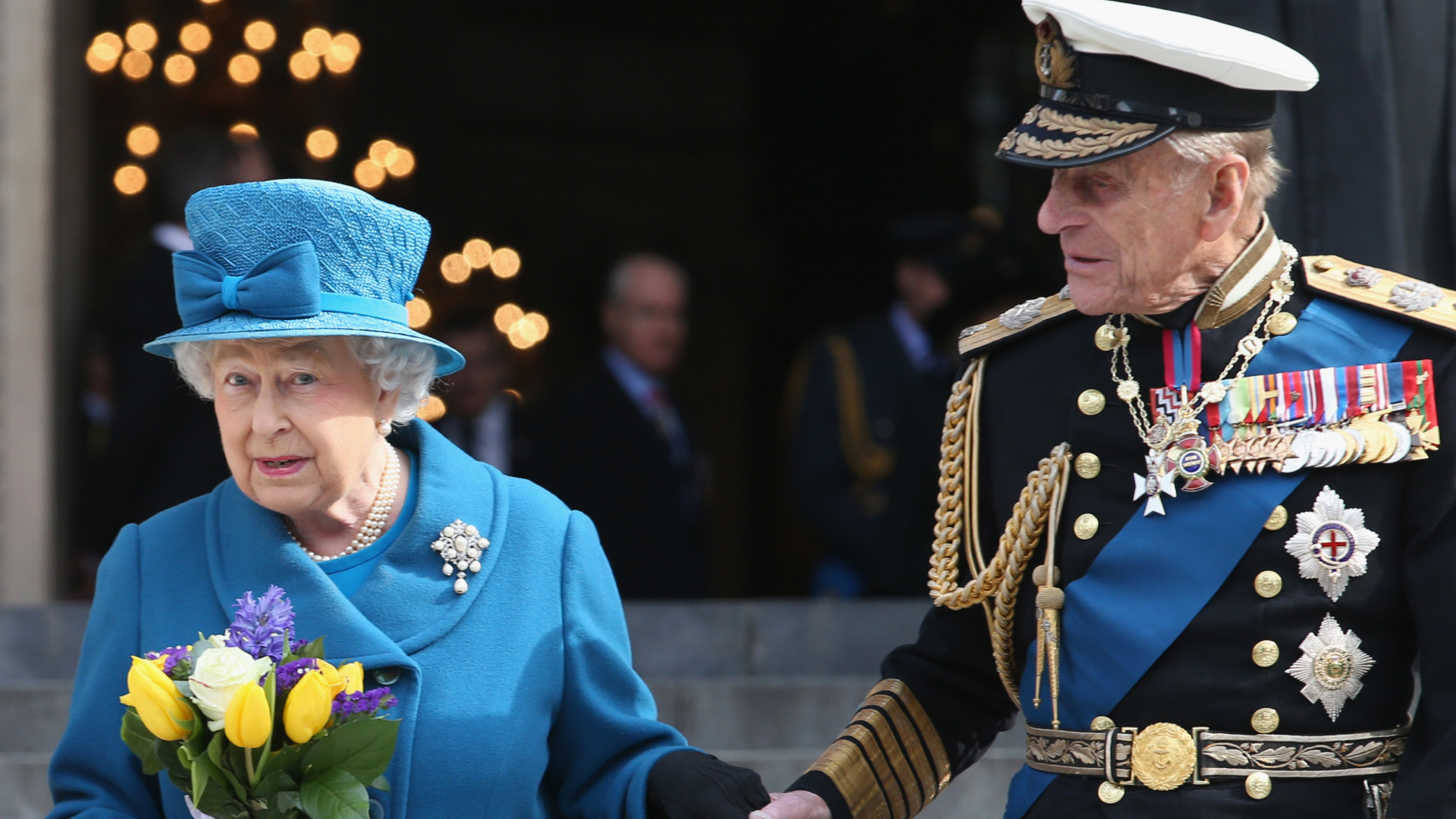 Queen Elizabeth wears a blue coat and matching hat and carries a bunch of tulips while her husband Prince Philip holds her hand