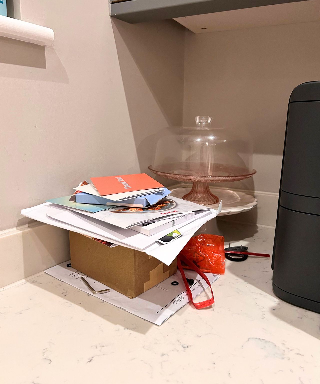 A box, piles of paperwork, cards and cake stands are positioned in the corner of a kitchen