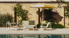 A seating area on a sun-drenched, off-white, stone pool deck. There is a seating area with a rope bench and matching rope arm chairs and a yellow and white parasol.