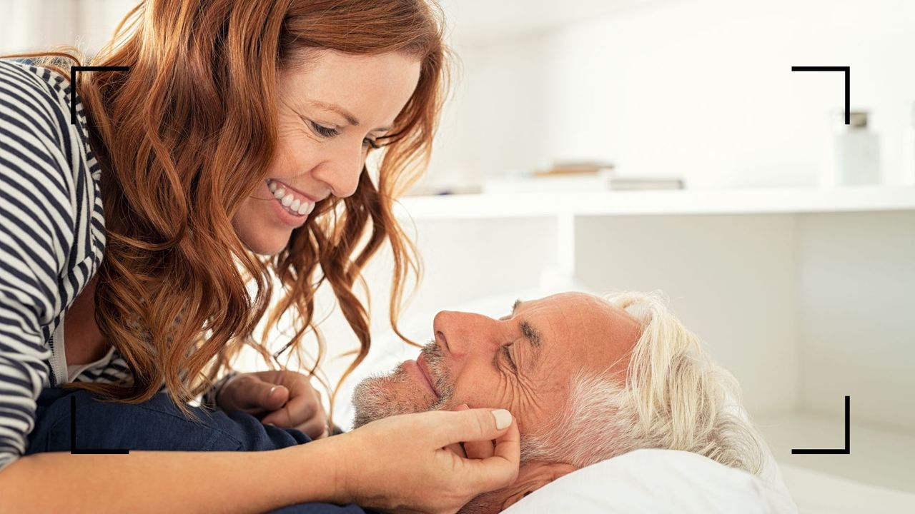 Woman lying on top of man in bed, smiling and laughing together with woman stroking man&#039;s face, representing the blooming orchid sex position