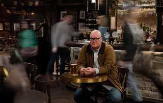 A troubled Paddy Kirk sits at a table as blurred people walk around him