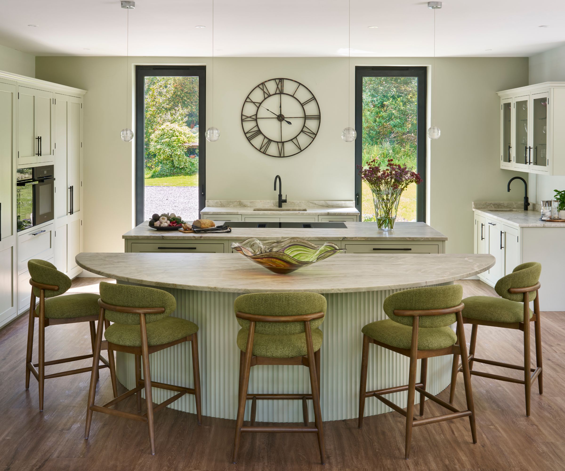 Light green kitchen with a curved island and ribbed base, topped with stone. Five green-upholstered stools face the island beneath glass pendant lights.