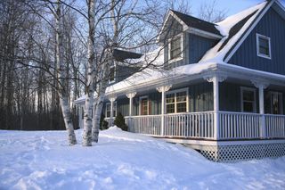 An older farm house with white veranda and two gables. At the front is a clump of birch trees. The snow is almost over the veranda.