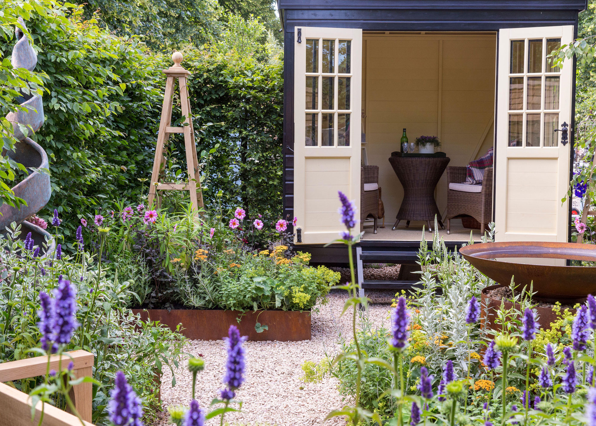raised garden shed with doors open, gravel path to the doors, flowerbeds with blooms and wooden obelisk