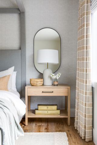 Bedroom with gray upholstered four poster gray wallcovering, wood nightstand with lamp and mirror above and wood floor and rug