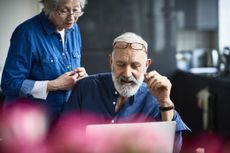 Senior man with beard using laptop and woman watching.
