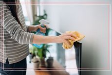 A woman wiping a TV with a cloth and holding a bottle of cleaning product