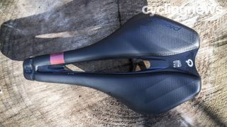 A top view of the Prologo Dimension AGX gravel saddle on a wooden surface, showing the large central cut out, ridged nose and carbon fibre shell between the padded flanks