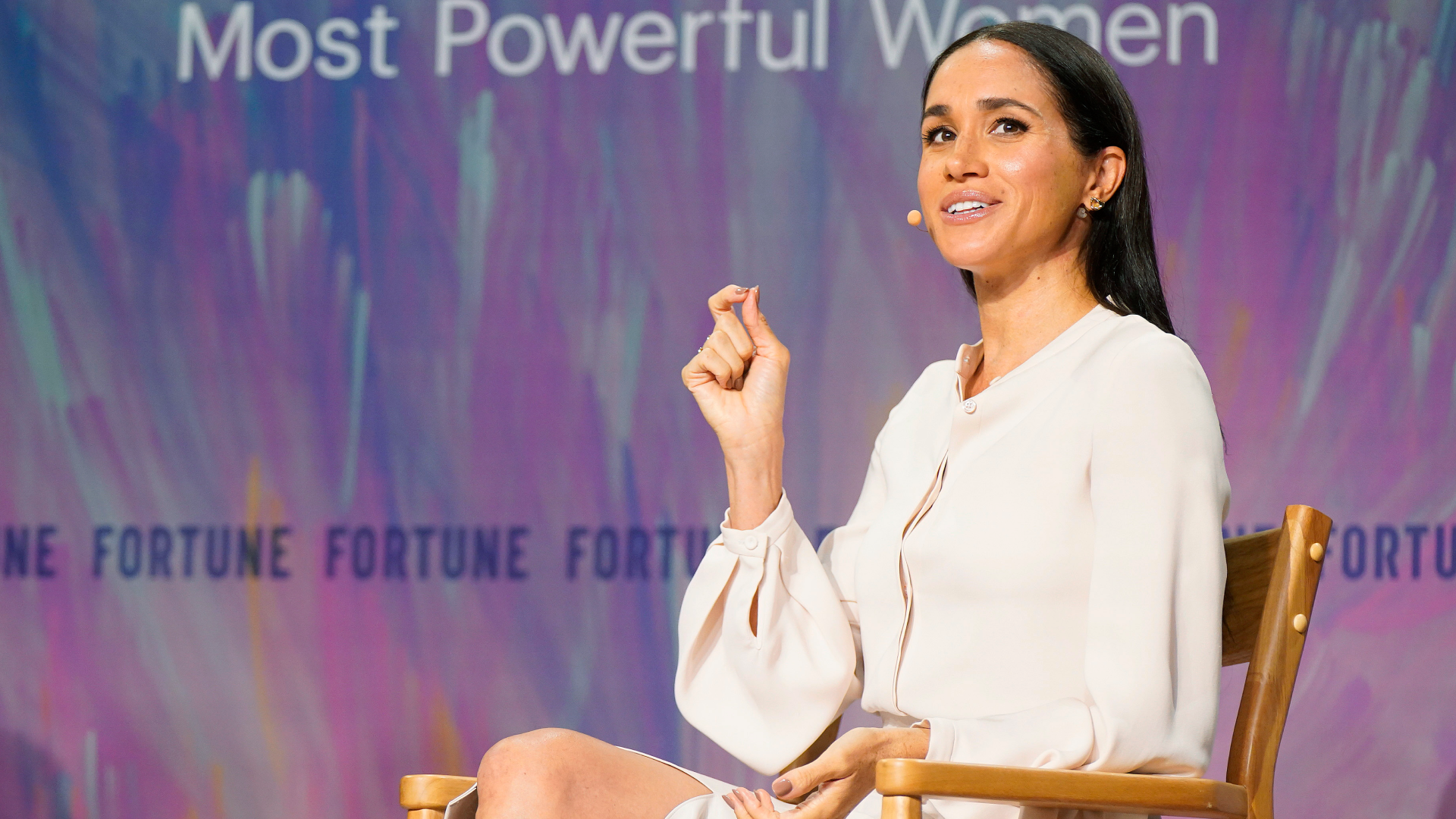 Meghan Markle wearing a white blouse sitting in front of a purple background