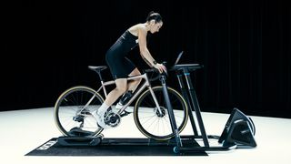 A white woman with dark hair rides a golden bike on a static trainer