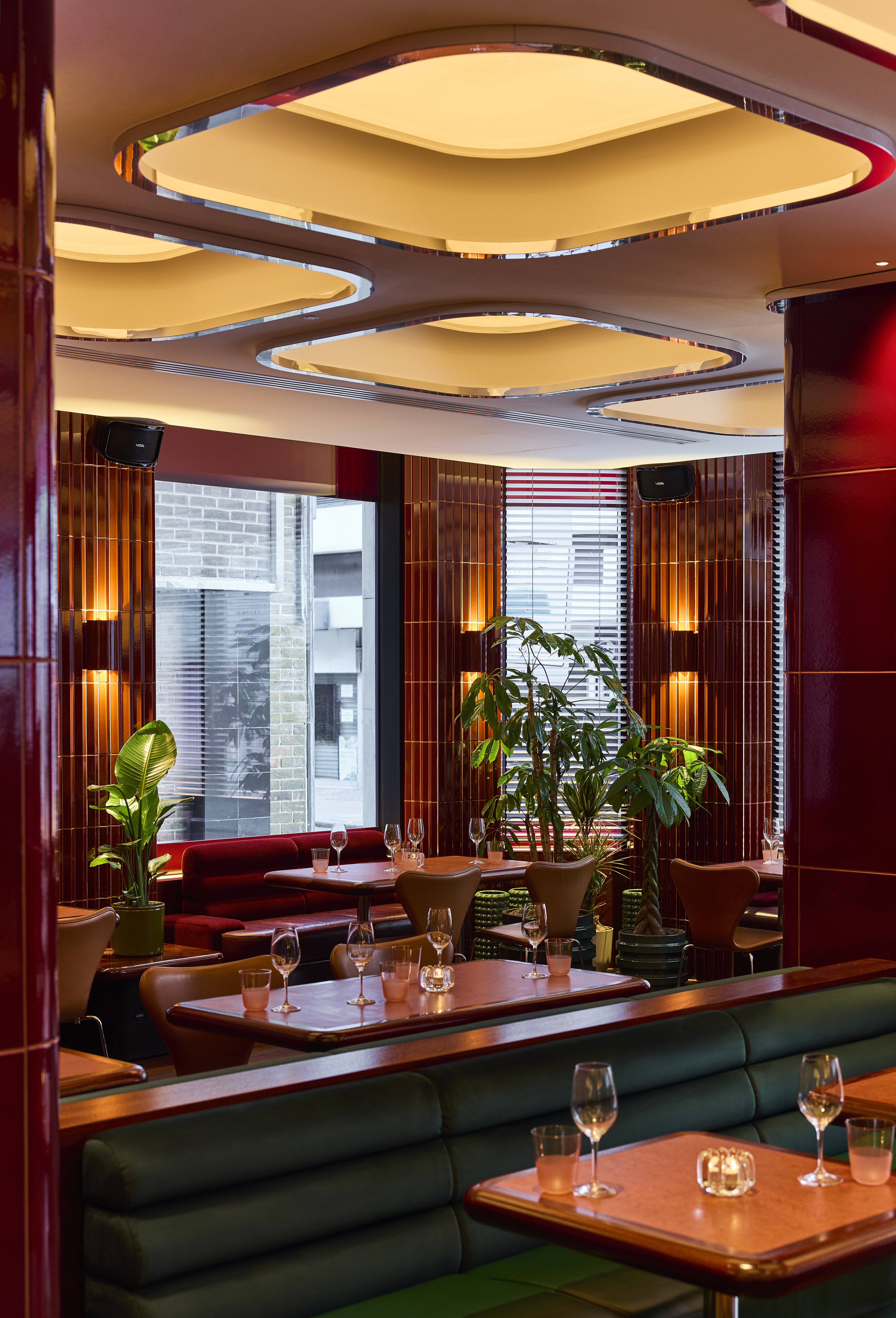The interiors of a fine dining restaurant feature coffered, back-lit ceilings with chrome detailing, cherry plum walls covered in glazed ceramic, dark green banquettes, and atmospheric sconces, as well as plants.