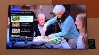 A Samsung TV on display at CES 2025. It is showcasing the AI-based Samsung Food software feature, which is correctly identifying a lasagna dish on a dinner table in the scripted content on screen. There is a smiling elderly woman cutting the lasagna, and a software-based pop-up on the screen detailing a list of lasagna recipes.