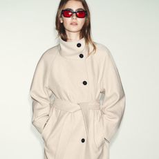 Model wears cream jacket with black buttons, red sunglasses
