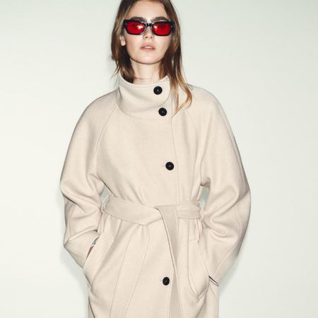 Model wears cream jacket with black buttons, red sunglasses