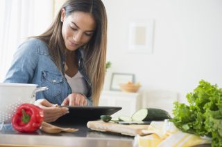A women sat at a work top surrounded by peppers, salad leaves and a chopped courgette on a chopping board. She is reading her digital tablet.