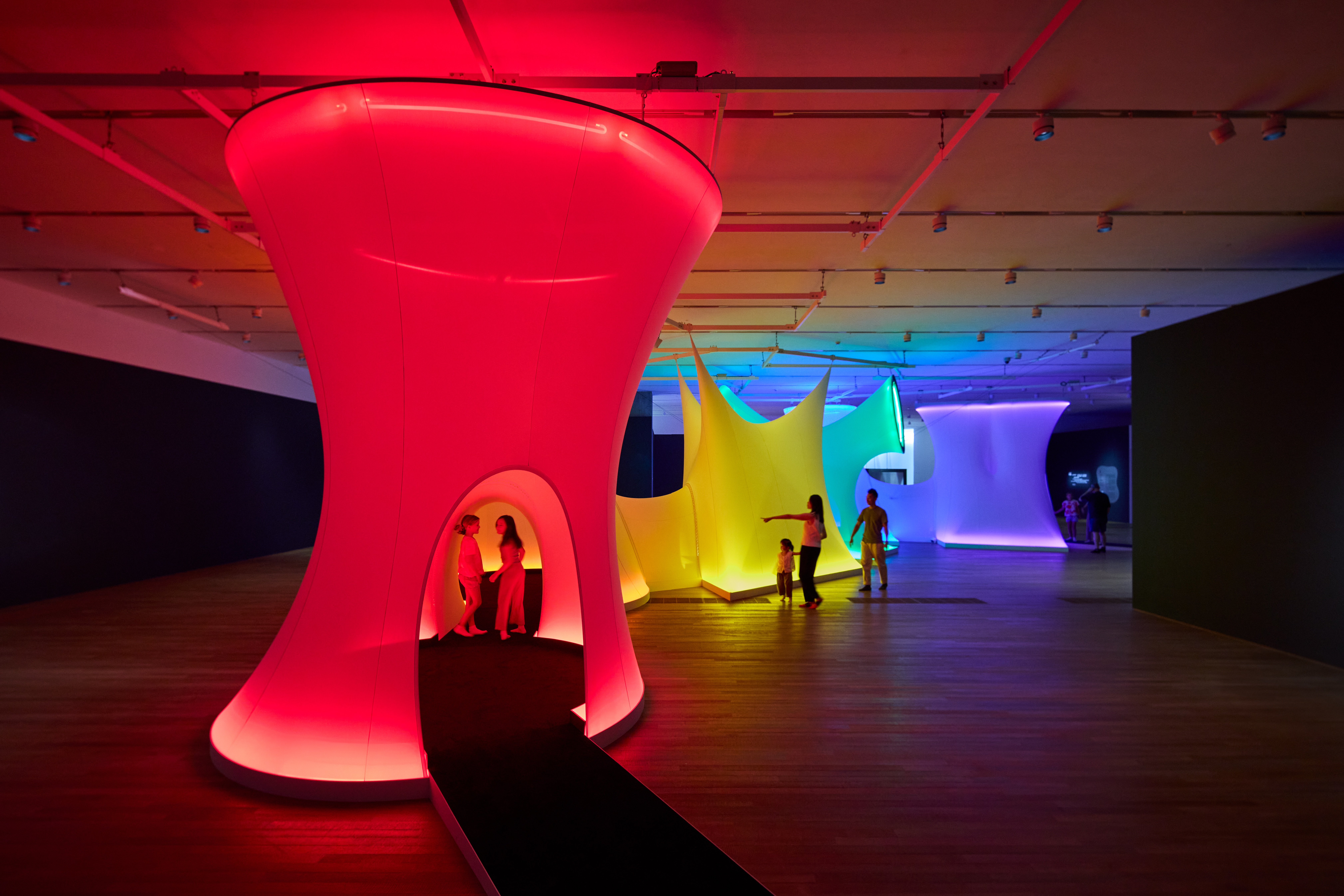 An immersive light installation in a gallery features multiple textile environments lit up in red, yellow, blue, and light green in which multiple adults and kids are playing and walking.