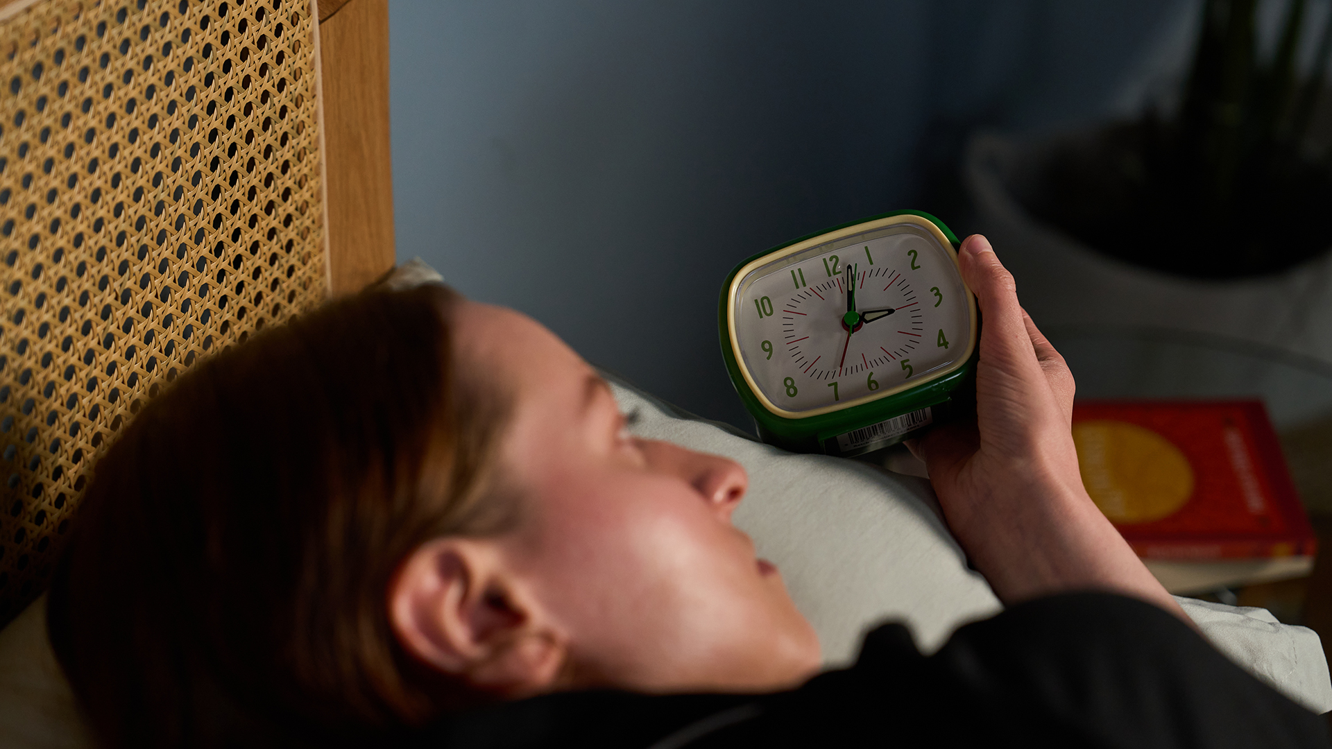 A woman lies in bed in the dark looking at an alarm clock she holds in her hand, showing the time is 3 a.m. 