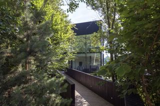 View of the wooden bridge that leads to the entryway. Trees surround the bridge, and we see a part of the all-glass villa.
