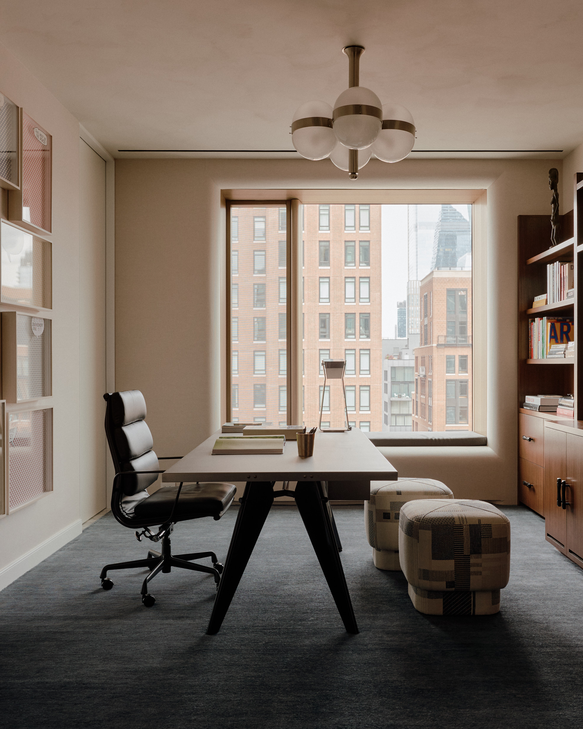 home office with dark gray carpets, big window, eames chair on desk floating in space with two ottoman, pendant light, bookshelf and frames on the wall