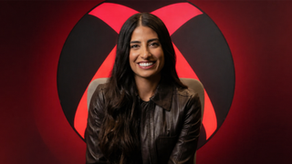 Asha Sharma sits facing the camera in a black leather jacket, positioned in front of a large Xbox-style logo, with the background and logo glowing in a red hue.