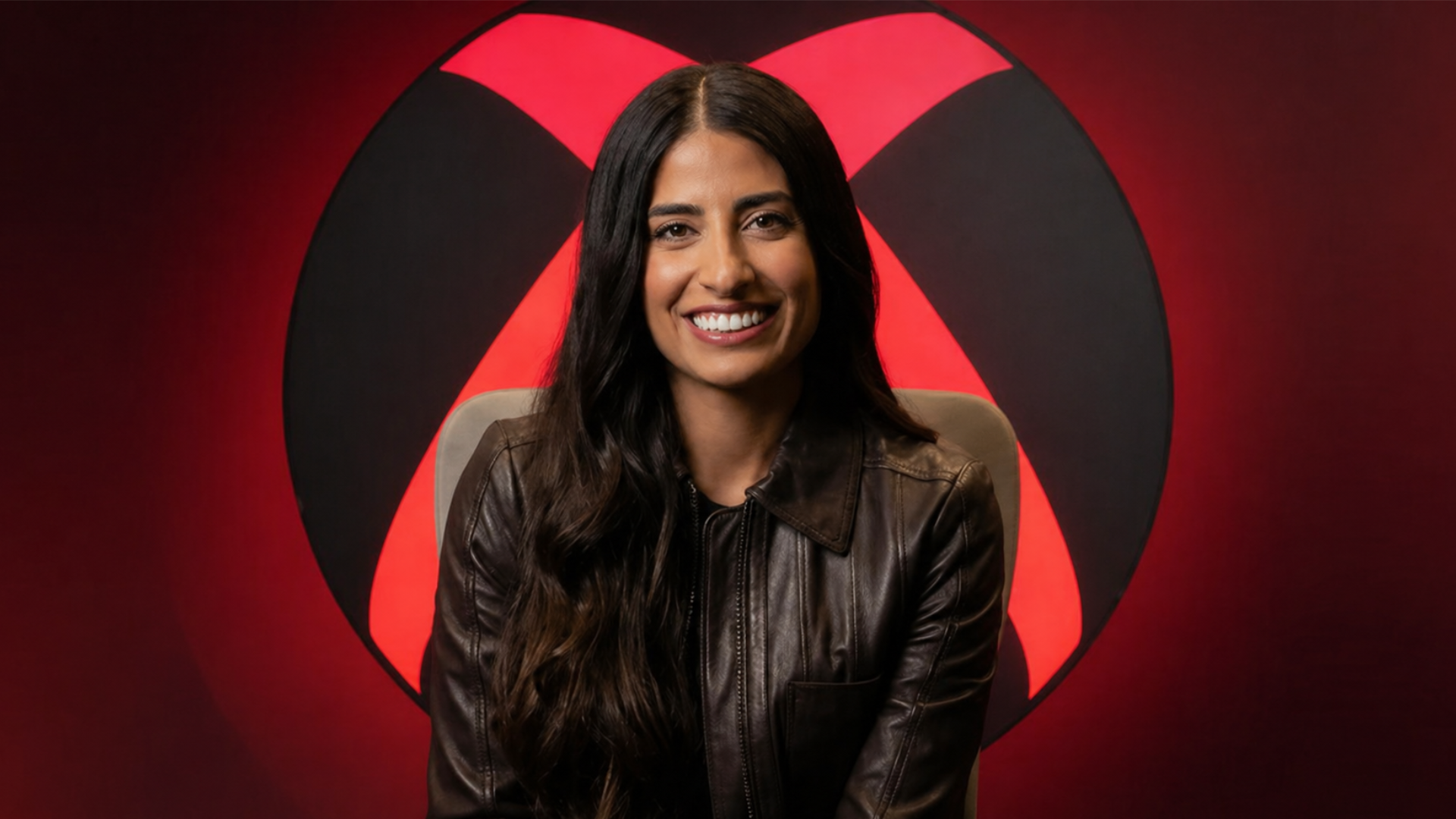 Asha Sharma sits facing the camera in a black leather jacket, positioned in front of a large Xbox-style logo, with the background and logo glowing in a red hue.