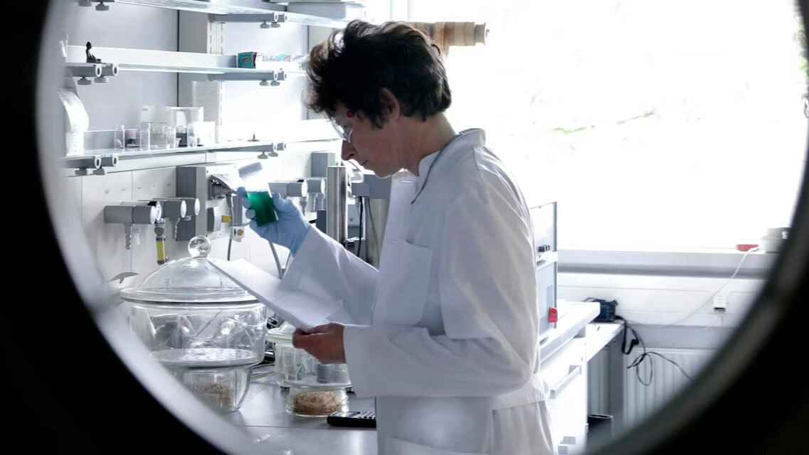 Photo of a scientist in a white lab coat looking at a device while holding a piece of paper. The scientist is facing left and leaning over a lab bench.