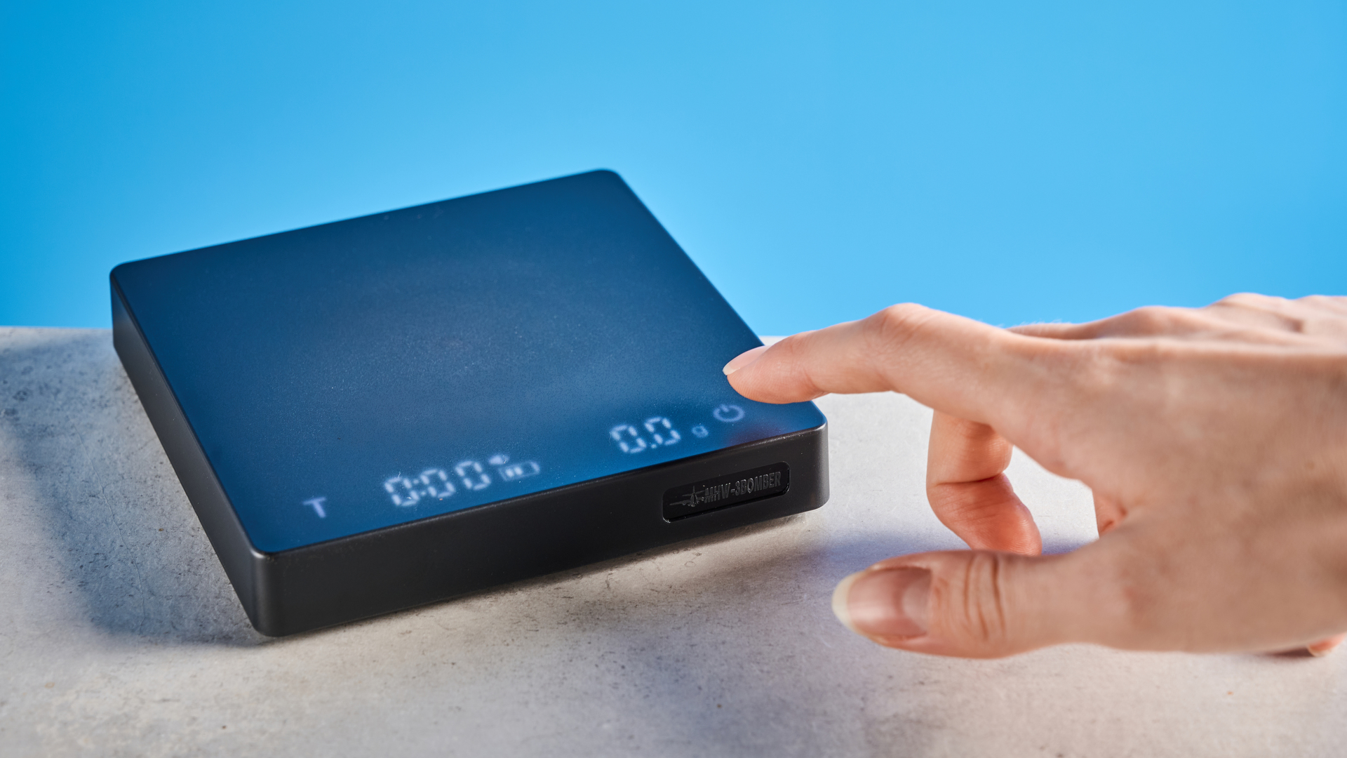 the mhw-3bomber cube coffee scale 2.0 in black photographed against a blue background showing its button controls and dial surface