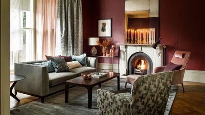 A cozy living room with burgundy walls, mismatched upholstered furniture, a large grey velvet rug, brown wooden coffee table, and fire burning in a stone fireplace with tall tapered candles burning on the mantelpiece above.