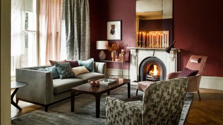 A cozy living room with burgundy walls, mismatched upholstered furniture, a large grey velvet rug, brown wooden coffee table, and fire burning in a stone fireplace with tall tapered candles burning on the mantelpiece above.