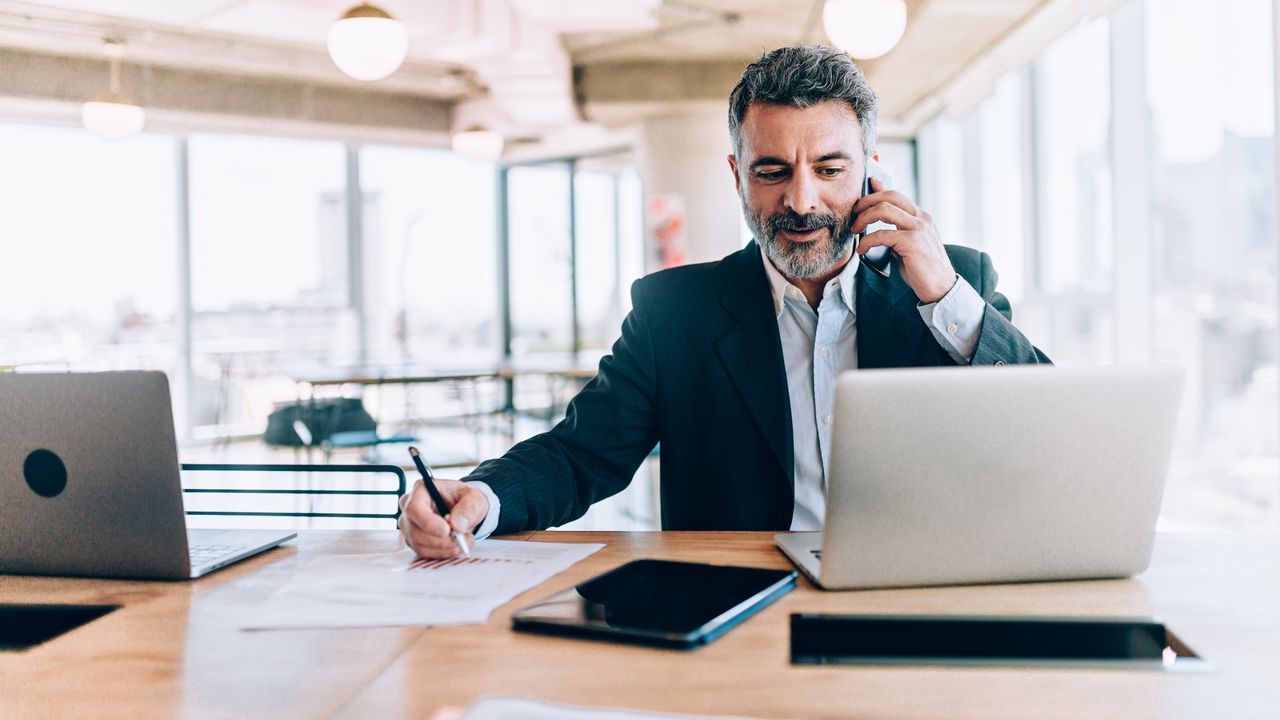 A businessman talks on his cellphone at his desk, taking notes.