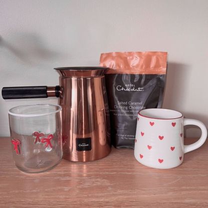 A Hotel Chocolat Velvetiser with a bag of salted caramel drinking chocolate, a white and red heart-patterned mug, and a glass with red bows. 