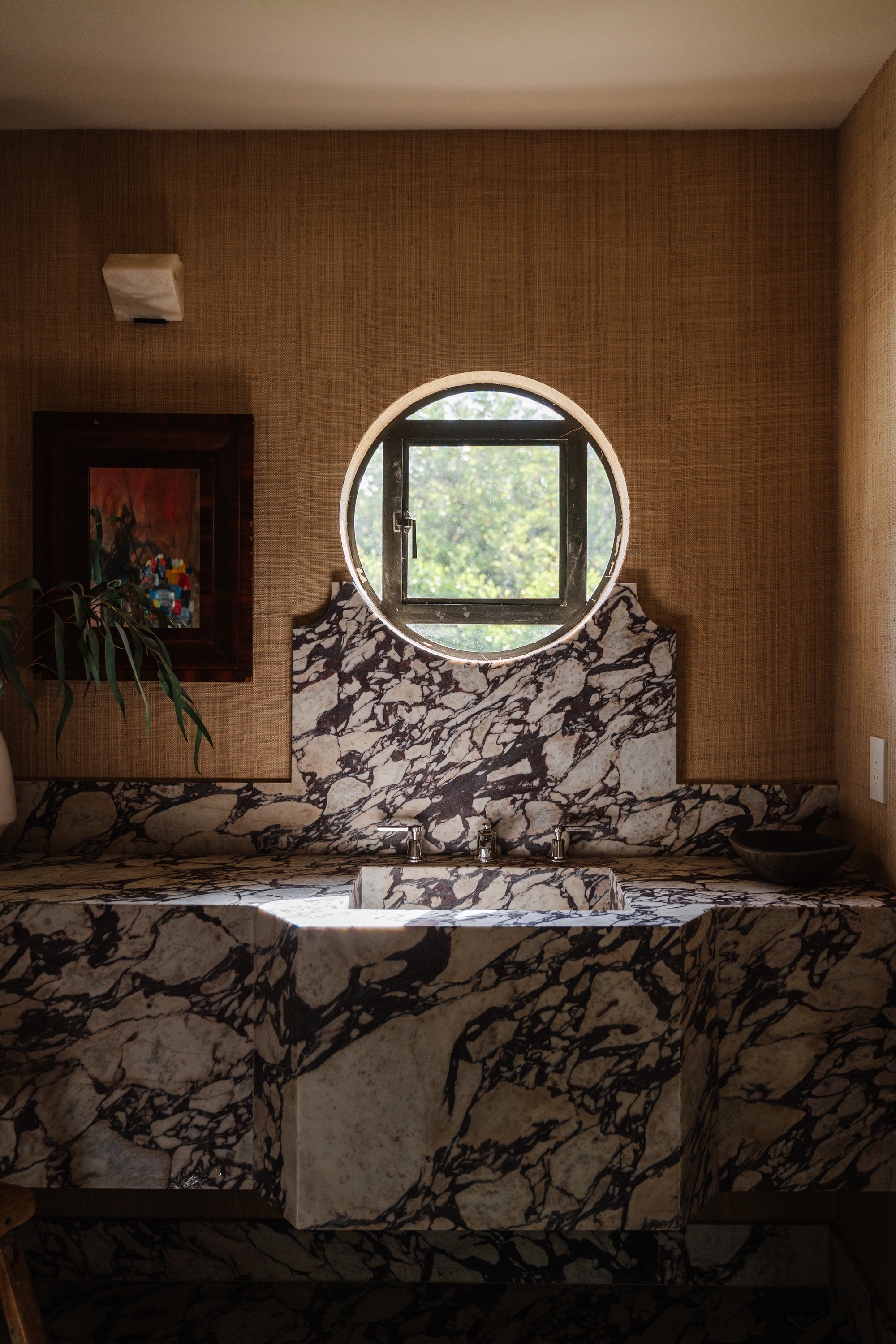 A powder room with glasscloth wall covering in a deep ocher tone with a dark brown-red veined marble sink with a round porthole window with a square black frame in the middle, a mirror and a plant