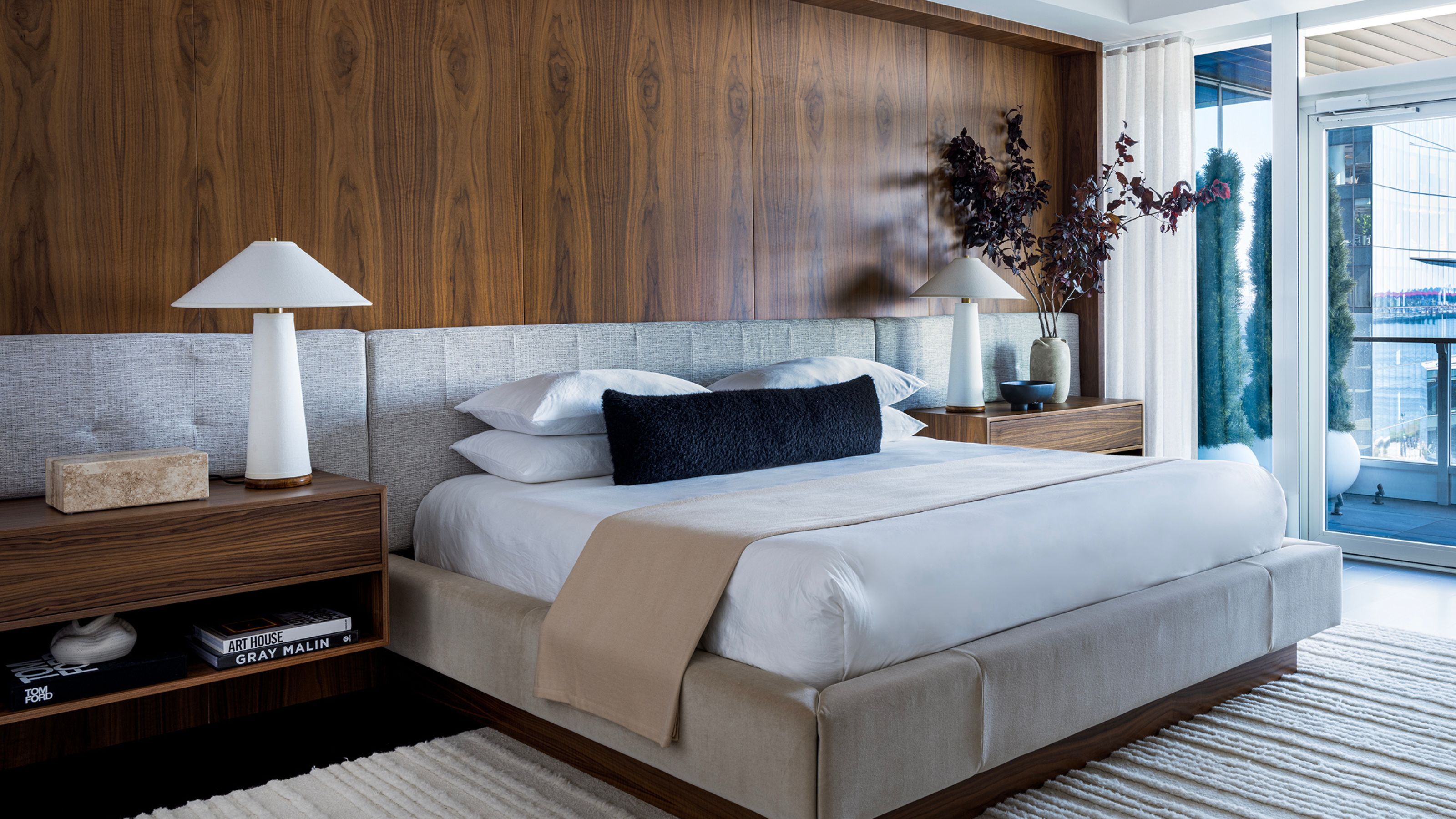 A large modern apartment bedroom with a wood paneled wall above the headboard, a large king-sized bed with white bedding, and a beige textured rug on the floor.