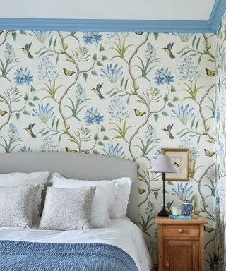 White wallpapered bedroom with wall patterns showing green long plants and blue flowers, gray headboard on a double bed with white, silver and blue bedding with scalloped edges