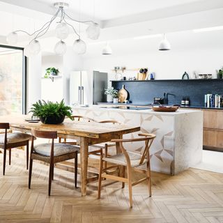 A white kitchen with herringbone-style wooden floor, wooden kitchen dining table, white tiled kitchen island, statement lighting fixture and navy splashback