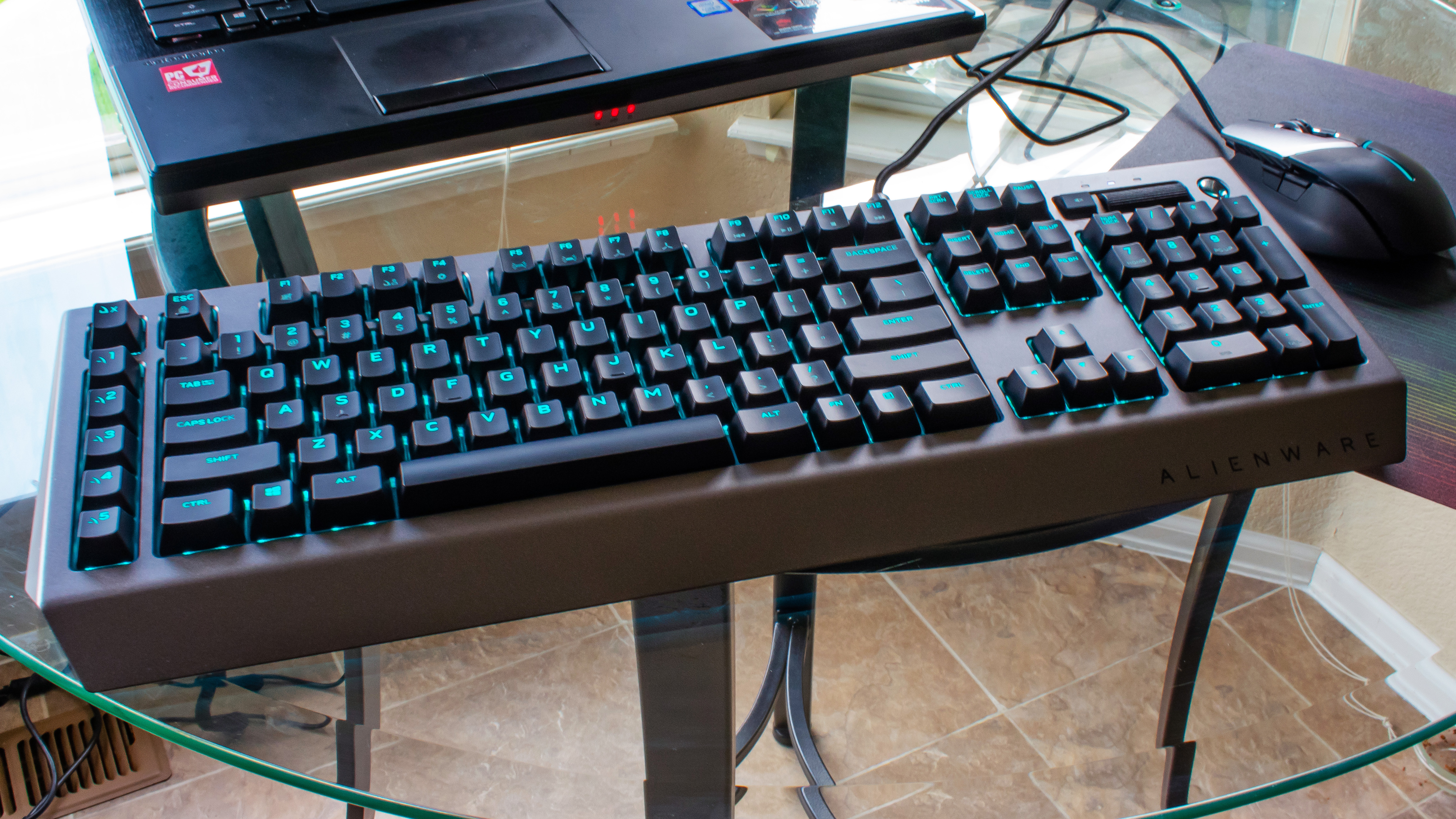 Alienware Pro Gaming Keyboard on a glass table next to a laptop and a mouse