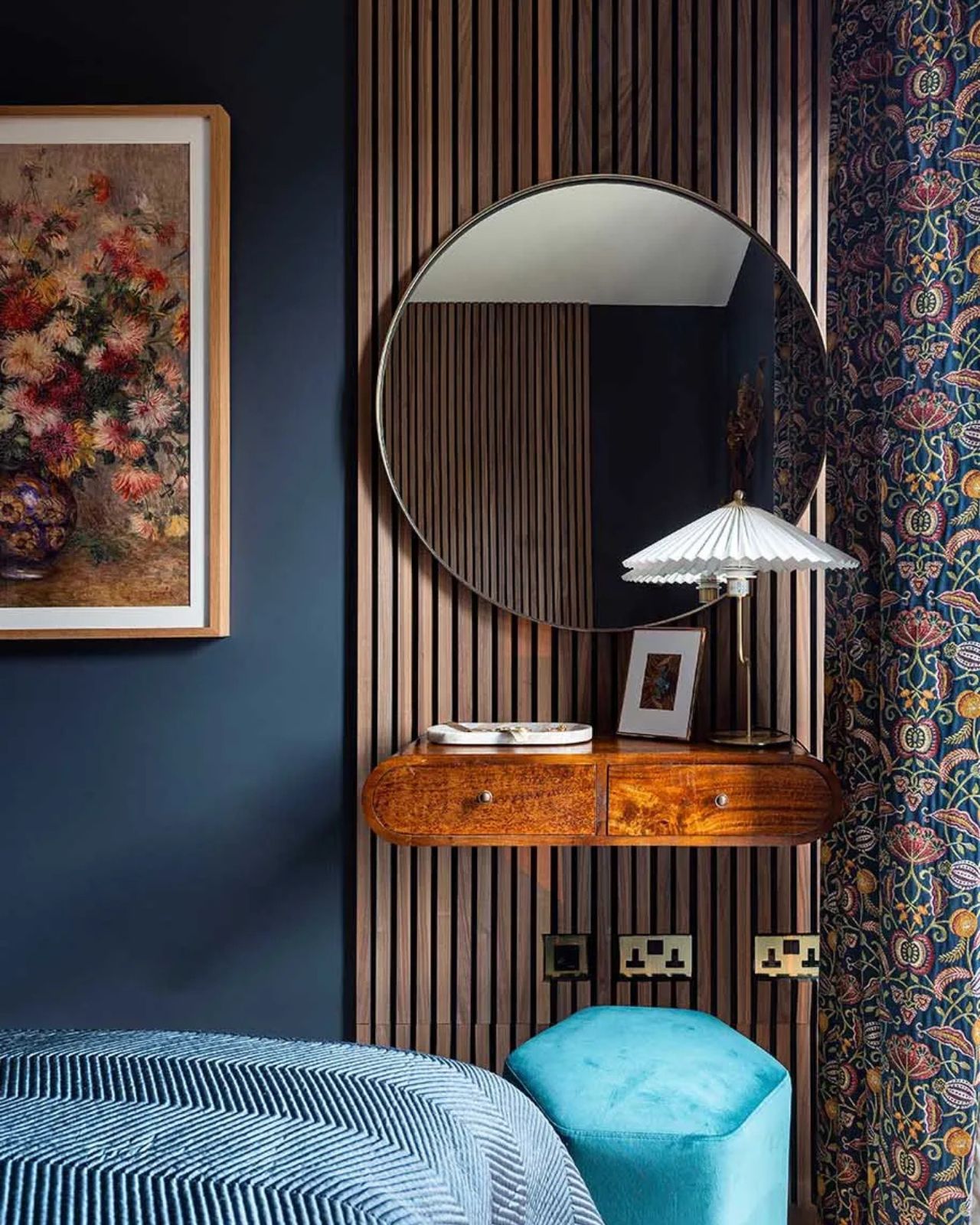 bedroom vanity floating on wall paneling, next to blue feature wall and artwork, with round mirror, floral curtains, and a lamp with a blue ottoman