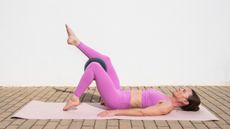 A woman practices Pilates toe taps on a mat outdoors. She is lying on the mat with her arms by her side and her knees bent; there is a Pilates ball between her knees. One foot taps the floor while the other is elevated. Behind her is a white-washed wall.