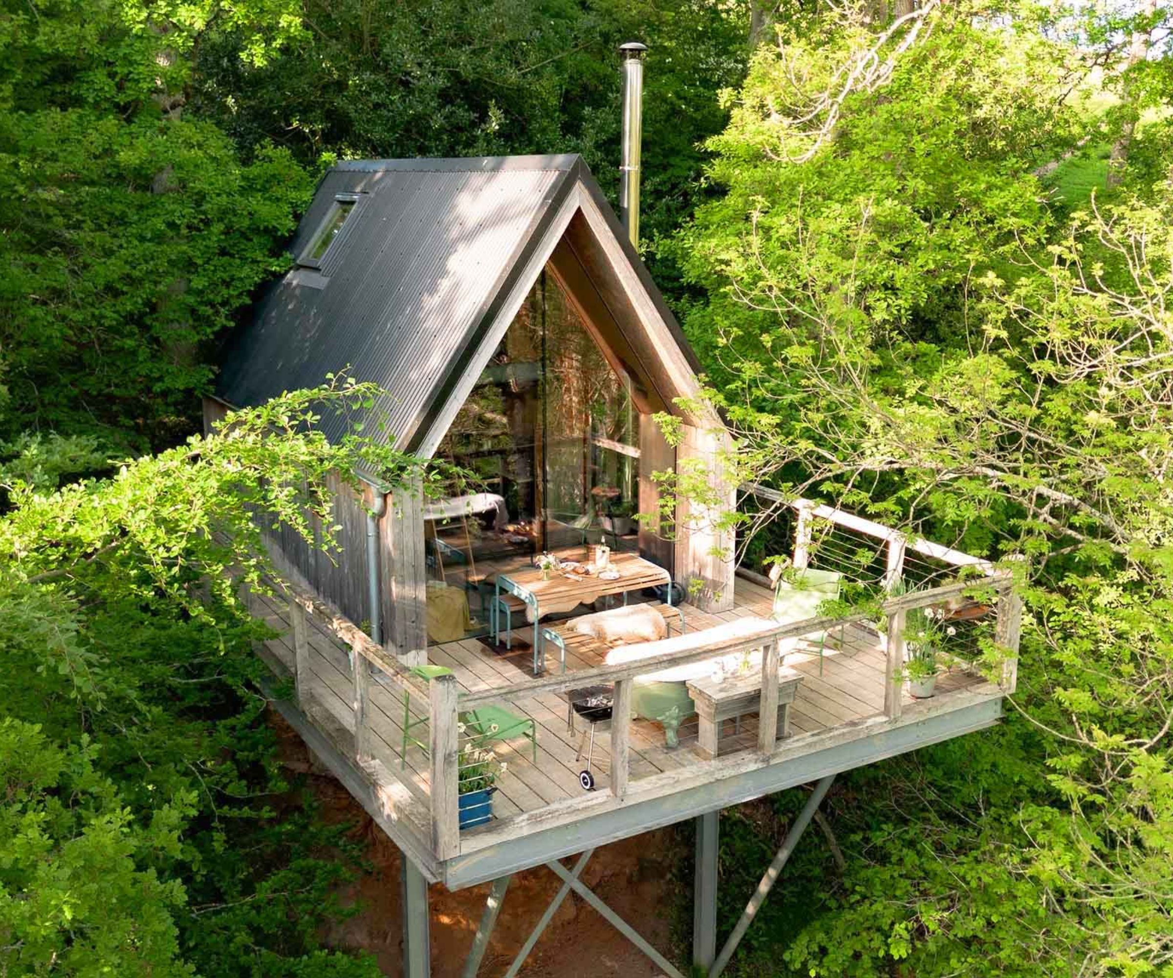 A modern treehouse cabin elevated on metal stilts, nestled high among leafy green trees. The compact A-frame structure features a steep black roof and full-height glass front, opening out onto a spacious wooden deck with outdoor seating and dining furniture.