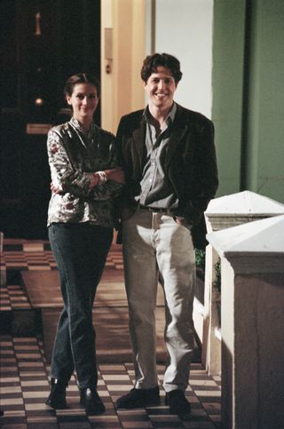 Julia Roberts wears blue jeans, black heeled boots, and a shirt standing next to Hugh Grant, who wears a brown jacket, a striped shirt, and linen pants on the set of the romantic comedy Notting Hill