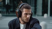 A man wears the Meze Strada headphones, which feature dark wood earcups and a metallic green frame, while looking to the side in an urban setting.