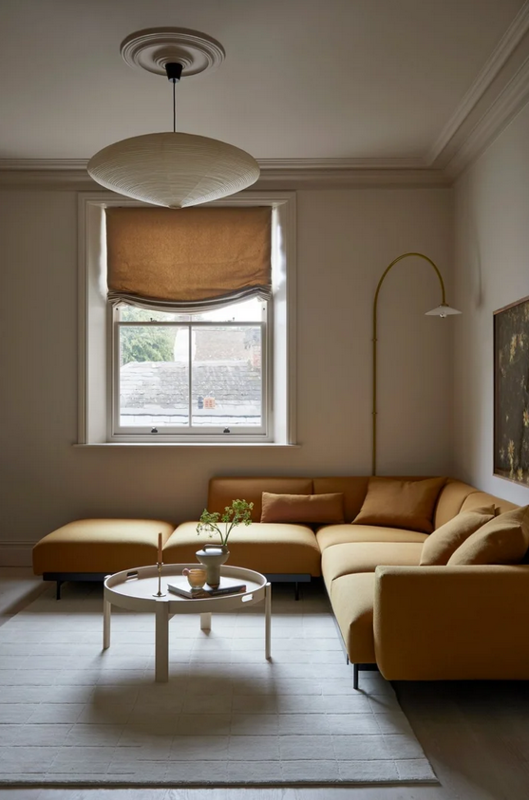 neutral living room with white rug, round coffee table styled with vase of flowers and book and candle stick, yellow corner sofa, wall lamp, a window with a relaxed roman blind and a pendant light