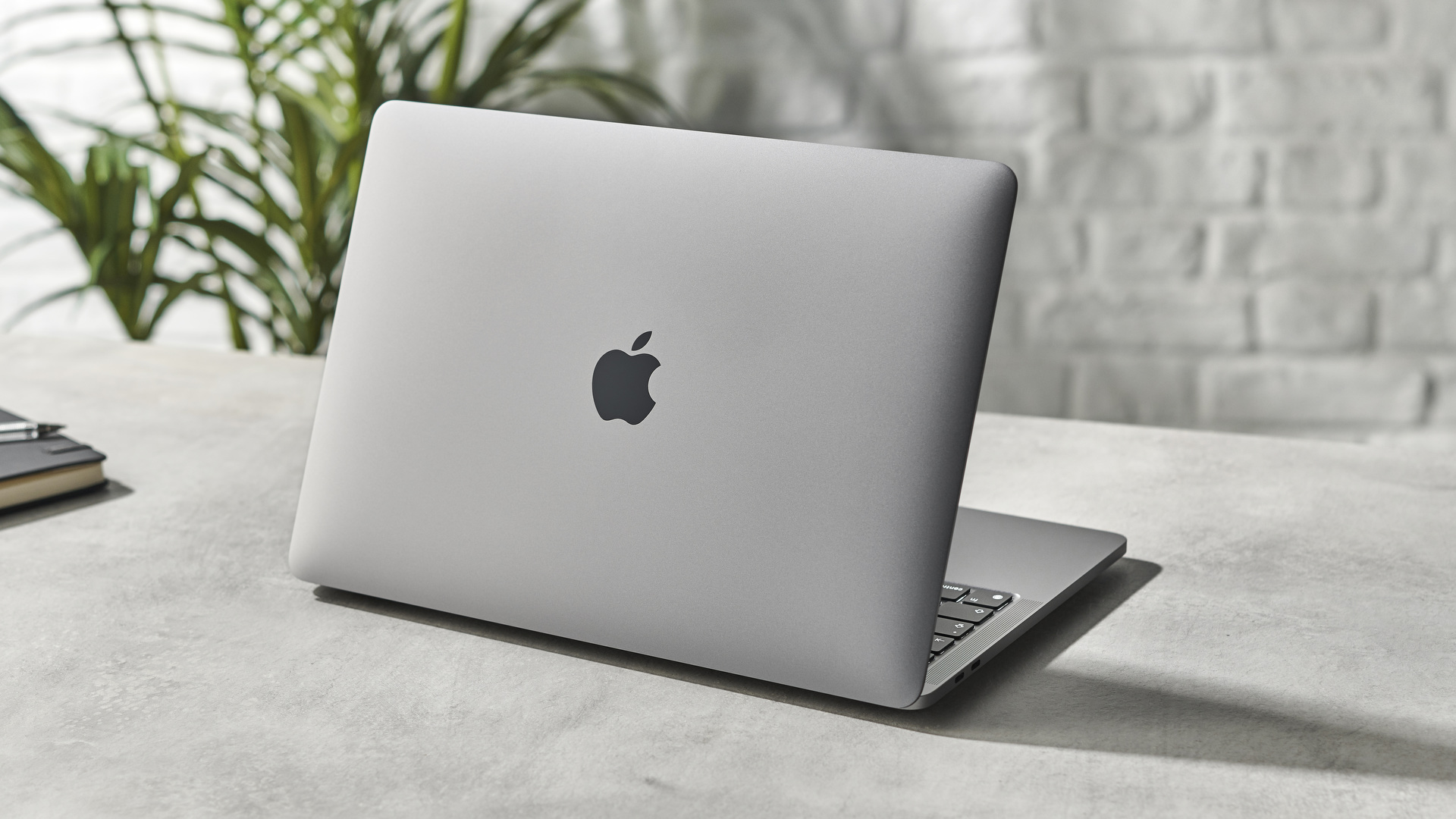 Apple MacBook Pro 13-inch (M1, 2020) on a desk next to a notebook in front of a white brick wall