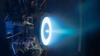 A solar electric propulsion thruster emits the blue hue of Xenon gas during testing. Vibrant blue light emanates in a circular shape from the dark grey thruster, which is mounted inside a vacuum chamber. The blue light then narrows into a plume as it moves farther away from the thruster, illuminating the otherwise darkened chamber.